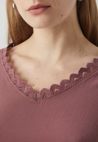 Lila felső, V-nyak körüli fodros csipke díszítéssel, texturált anyagból készült, laza szabással, amely simán esik.