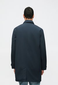 Muž v tmavomodrom dlhom kabáte stojí chrbtom k fotoaparátu na jednolatom svetlom pozadí.