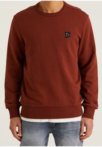 Sweat-shirt couleur rouille avec texture côtelée, col rond et patch logo noir. Équipé de poignets et d'un ourlet ajustés. Porté sur une chemise blanche.