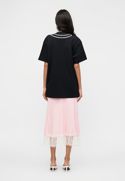 Μαύρο υπερμεγέθες T-shirt με διακοσμητική λευκή πέρλα στη λαιμόκοψη, συνδυασμένο με ροζ σατέν φούστα με λευκή δαντέλα στο τελείωμα.