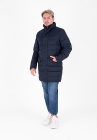 Piumino blu navy con colletto alto, chiusura a bottoni e zip, due tasche frontali e una finitura testurizzata, abbinato a jeans blu e sneakers bianche.