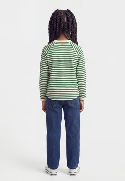 Bambino con capelli lunghi e intrecciati che indossa una maglia a maniche lunghe a righe verdi e bianche, jeans blu e scarpe da ginnastica bianche, visto di schiena.