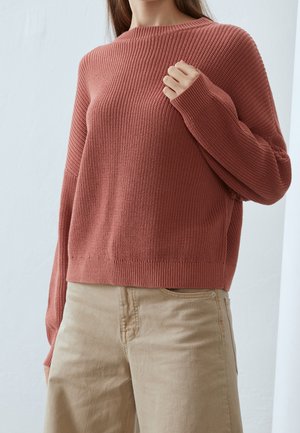Femme portant un pull en maille côtelée rose poussiéreux et un pantalon beige, une main agrippant légèrement le pull près du col.