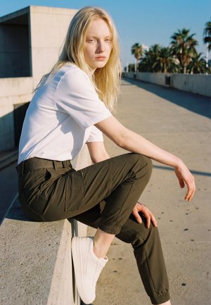 Une femme blonde, vêtue d’un T-shirt blanc, d’un pantalon foncé et de baskets blanches, est assise sur un muret en béton à côté d’une allée bordée de palmiers.