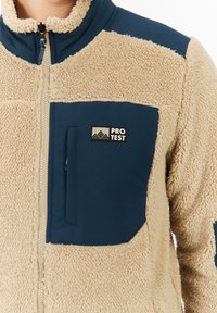 Beige fleecová bunda s námořnickomodrou kapsou na hrudi, vysokým límcem, zipovým uzávěrem a logem "Pro Test" na kapse.