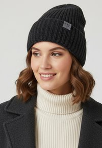 Black ribbed beanie with a folded cuff and logo patch. Worn with a light-colored ribbed turtleneck and dark coat, highlighting texture and fit.