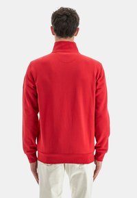 Felpa rossa con colletto alto, maniche lunghe, orlo a coste e texture liscia. Design minimalista, senza loghi o motivi visibili.