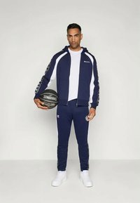 Ναυτικό αθλητικό κουστούμι φτιαγμένο από μαλακό ύφασμα; ζακέτα με φερμουάρ και λευκές λεπτομέρειες, ταιριαστά παντελόνια και μία μαύρη μπάλα μπάσκετ. Λευκά αθλητικά παπούτσια.