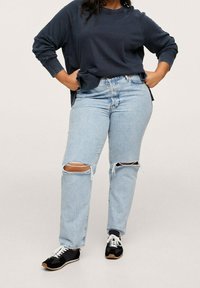 Jeans blu chiaro strappati con buchi sulle ginocchia, abbinati a un maglione navy. Scarpe nere con lacci bianchi e suola color beige. Look e stile casual.