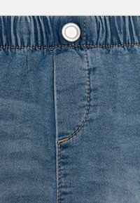 Shorts en denim de couleur bleu clair, avec une taille élastique, un bouton-pression argenté et un surfilage contrastant de couleur beige sur les coutures et la poche.