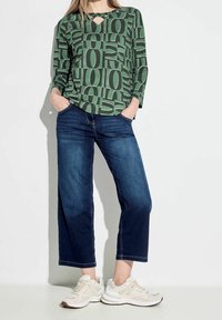 Top lungo a maniche lunghe verde con motivo e dettaglio a chiave, abbinato a jeans a gamba larga blu e sneakers beige. Tessuto morbido, vestibilità casual.