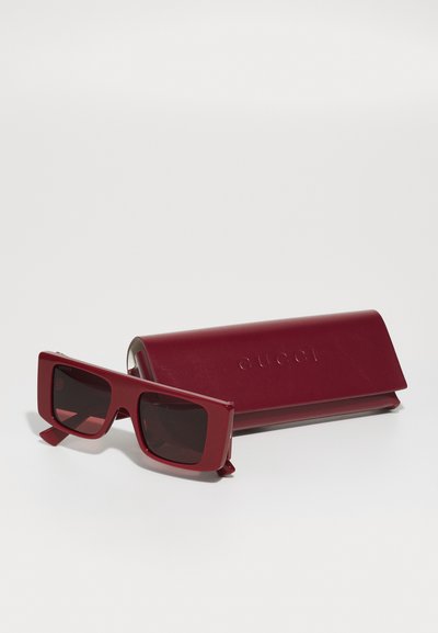 Ochelari de soare dreptunghiulari roșii, cu lentile întunecate, având o ramă groasă. Însoțiți de un toc din piele roșie asortat, gravat cu "GUCCI."