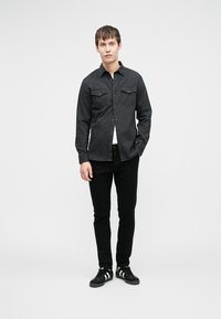 Camicetta nera con bottoni e due tasche sul petto, abbinata a jeans neri e sneakers nere con strisce bianche. Design minimalista, senza motivi.