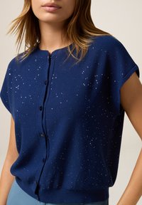 Eine Frau trägt ein kurzärmliges, dunkelblaues Hemd mit glitzernden weißen Punkten, kombiniert mit hellblauen Hosen, vor einem schlichten Hintergrund.
