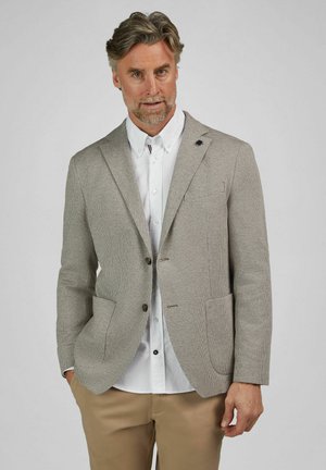 Mann, der einen beigen karierten Blazer über einem weißen Hemd und braunen Hosen trägt, steht mit einer Hand in der Tasche vor einfachem Hintergrund.