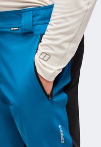 Main dans la poche zippée d’un pantalon d’extérieur bleu et noir, portée avec un haut beige clair à manches longues et petit logo sur la manche.