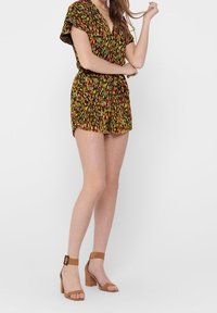 Kwiatowy kombinezon z krótkimi rękawami, dekoltem w serek, podkreśloną talią i szortami. Brązowe sandały na słupku z paskami na kostce. Wzór w kolorach zielonym i pomarańczowym.