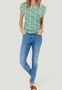 Top de manga corta con estampado floral verde, combinado con jeans ajustados de color azul claro. Los zapatos son sandalias marrones con tiras, con un estilo casual.