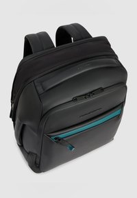 Mochila negra con correas acolchadas, múltiples compartimentos con cremallera, incluido uno con tirador de cremallera en color verde azulado, y un panel frontal de textura suave.