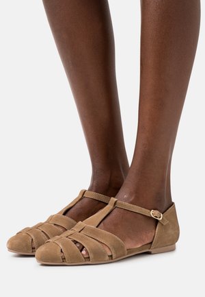 Ballerines en suède beige avec un design à T, featuring des lanières croisées, un bout arrondi et une boucle à la cheville pour un maintien sécurisé. Texture douce, profil minimal.