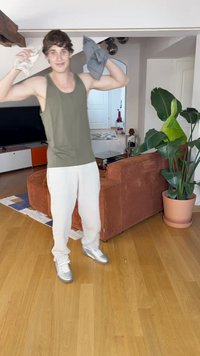 Un débardeur vert olive, un pantalon blanc et des baskets argentées. Tenant un objet gris dans une main et un objet beige dans l'autre, dans un cadre intérieur.