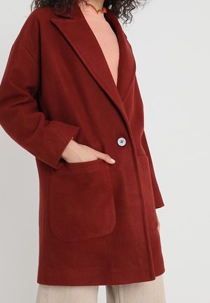 Persoon draagt een lange roestkleurige jas met grote voorzakken, één hand in een zak, over een lichtroze geribde top en een beige broek.