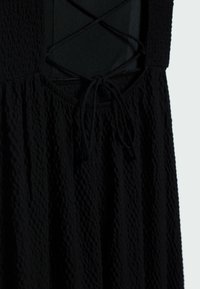 Robe en tissu noir texturé avec un détail en dentelle croisée et un nœud noué à la taille, montrant un gros plan de la partie supérieure et du milieu du corps.