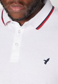 Biała koszulka polo z fakturowanego materiału, z kołnierzykiem w czerwono-granatowe paski, trzema guzikami i małym, ciemnoniebieskim logo w kształcie ptaka.