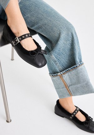 Zapatos planos negros con un cuello de pelusa negra, correa ajustable con herrajes plateados, combinados con jeans de pierna ancha azul claro.