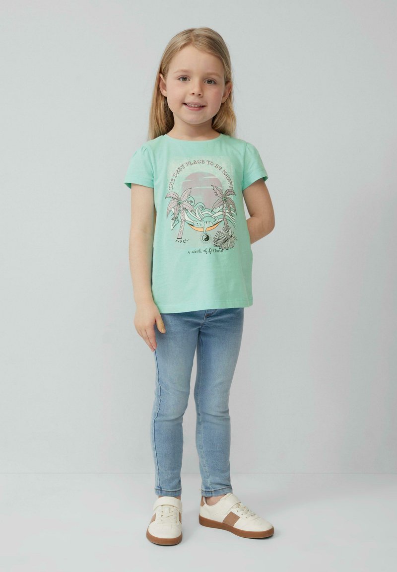Jong meisje met blond haar, gekleed in een mintgroene graphic t-shirt, lichte blauwe jeans en witte sneakers, staand tegen een effen achtergrond.