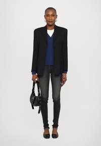 Black blazer with side ruching, layered over a blue cardigan, paired with dark jeans and black flats. Accessories include a small black bag.