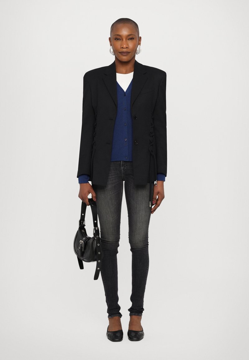Black blazer with side ruching, layered over a blue cardigan, paired with dark jeans and black flats. Accessories include a small black bag.