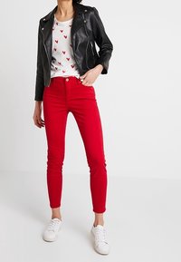 Czarna skórzana kurtka motocyklowa na białej koszulce z czerwonymi wzorami serc, w połączeniu z czerwonymi obcisłymi dżinsami i białymi trampkami.