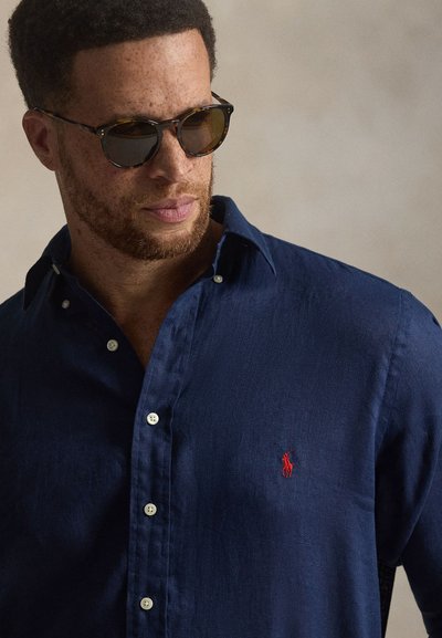 Rövid, göndör hajú és szakállas férfi, aki teknőcpáncél mintás napszemüveget és tengerészkék gombos inget visel piros hímzett logóval.