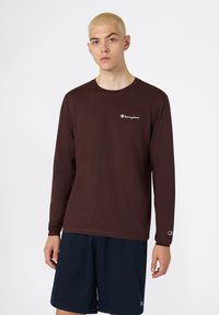 T-shirt brun à manches longues en coton, avec un petit logo blanc sur la poitrine gauche et un col rond, associé à un short marine.