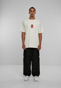 Kremfarget oversized t-skjorte med kort erme og et grafisk trykk på forsiden; kombinert med løse svarte cargo-bukser og hvite sneakers.