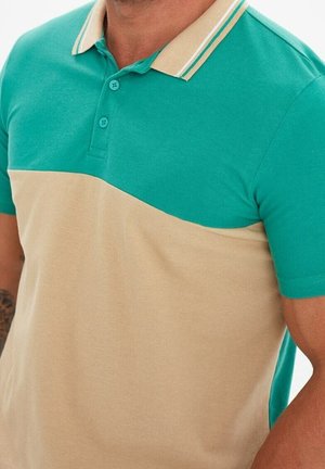 Man draagt een poloshirt met korte mouwen met een turquoise bovenstuk en een beige onderstuk, met een beige kraag met turquoise strepen.