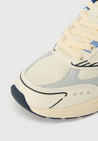 Tenis bej cu plasă și suprapuneri sintetice, accente gri, șireturi crem și detalii negre și albastre pe partea superioară. Talpă din cauciuc rotunjită.