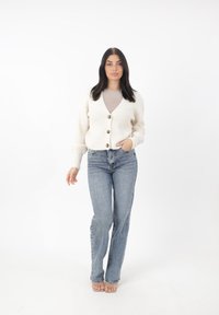 Cardigan lavorato a maglia color avorio con scollo a V e tre bottoni marroni, abbinato a jeans svasati azzurri chiari e sandali trasparenti.