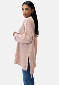 Hellrosa gestrickter Pullover in lockerer Passform mit langen Ärmeln, seitlichen Schlitzen und strukturiertem Stoff. Kombiniert mit blauen Jeans, seitlich abgebildet.