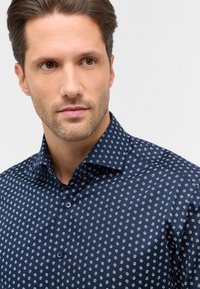 Camicia navy con bottoni, caratterizzata da piccoli motivi geometrici bianchi. Presenta un colletto a punta e un tessuto texturizzato. Due bottoni visibili sulla pattina.