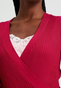 Fuchsiový žebrovaný cardigan s výstřihem do V, vyrobený z texturovaného pleteného materiálu, vrstvící se přes bílou krajkovou blůzu s vlnitým okrajem.