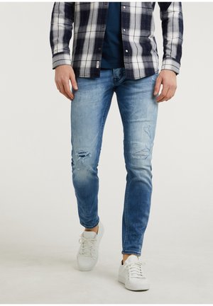 Man draagt lichtblauwe ingercheurde skinny jeans, witte sneakers en een zwart-wit geruit shirt en loopt naar voren.