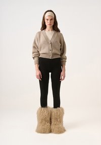 Cardigan tricotat bej cu butoni, colanți negri și cizme pufoase bej din blană; textură fină, croială lejeră și design stratificat.