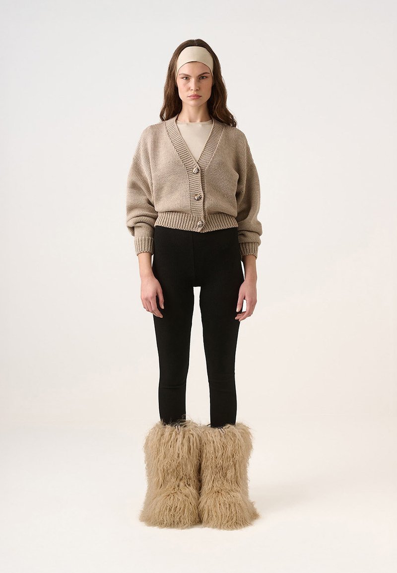 Cardigan tricotat bej cu butoni, colanți negri și cizme pufoase bej din blană; textură fină, croială lejeră și design stratificat.