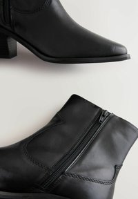 Botas de cuero negro hasta el tobillo con punta afilada, cremalleras laterales y tacón apilado. Presentan una textura suave y detalles de costura en contraste.