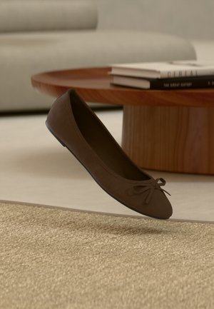 Ballerine en daim marron avec un petit nœud sur le bout, placée près d'une table basse ronde en bois avec des livres dessus, sur un tapis beige.