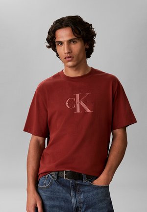 Ung mand med krøllet mørkt hår iført en vinrød Calvin Klein t-shirt og blå jeans med den ene hånd i lommen mod en grå baggrund.