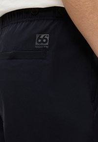 Gros plan sur un pantalon bleu marine foncé avec une poche arrière et le logo « 66° North » près de la ceinture, porté par une personne en chemise blanche.