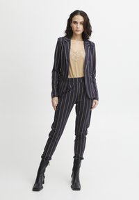 Marineblauer Nadelstreifenanzug mit tailliertem Blazer und schmal geschnittenen Hosen, kombiniert mit einem beigen Oberteil mit Spitzenbesatz und schwarzen Absatzstiefeln. Glatte Textur.